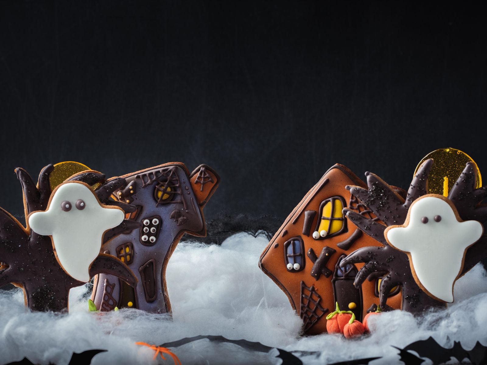 two flat gingerbread house cookies with chocolate icing and white ghost cookies standing in cotton wool fog