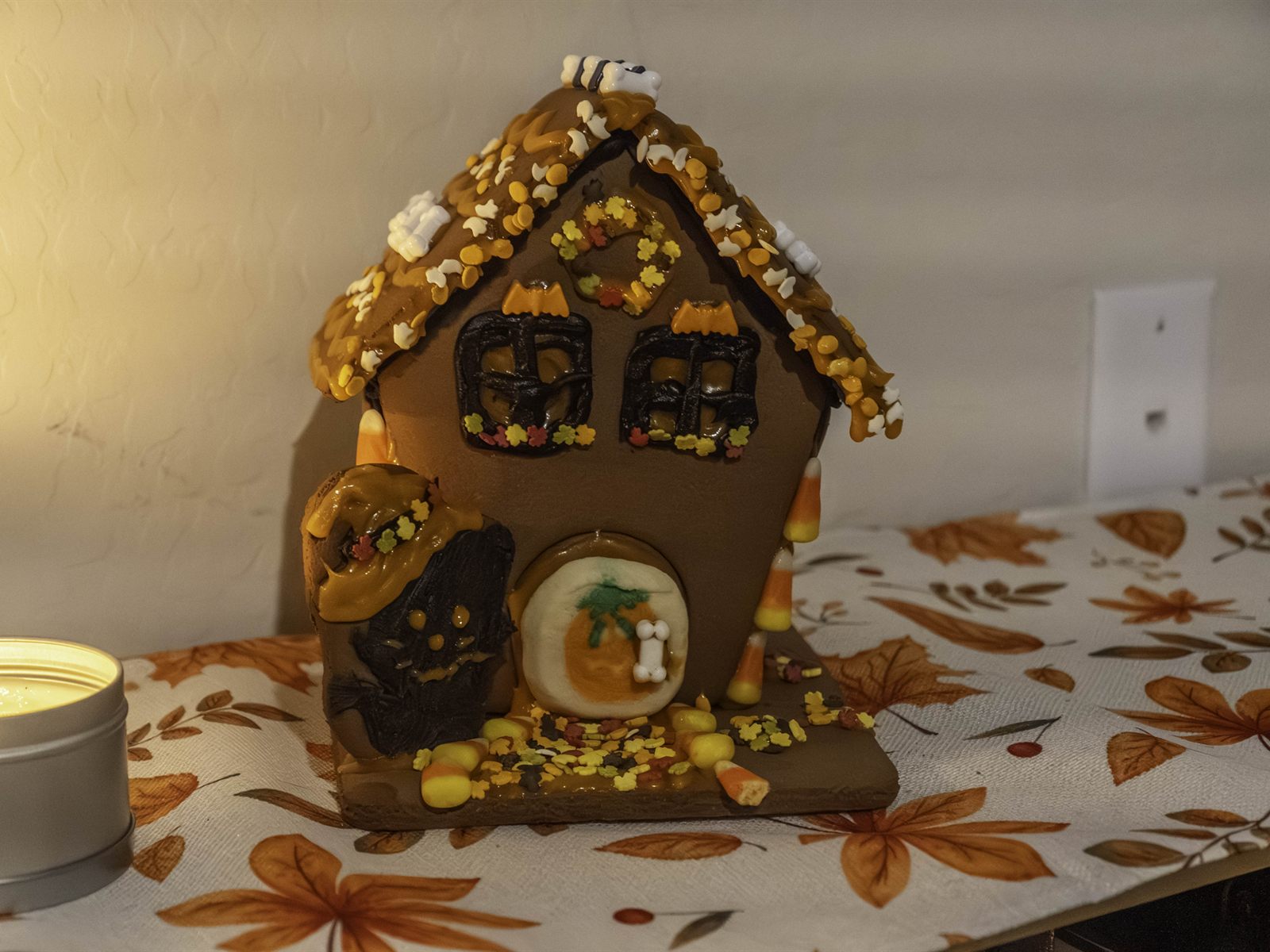 homemade gingerbread house with an orange icing roof, autumn sprinkles and candy corn, lit by a candle on a fall tablecloth