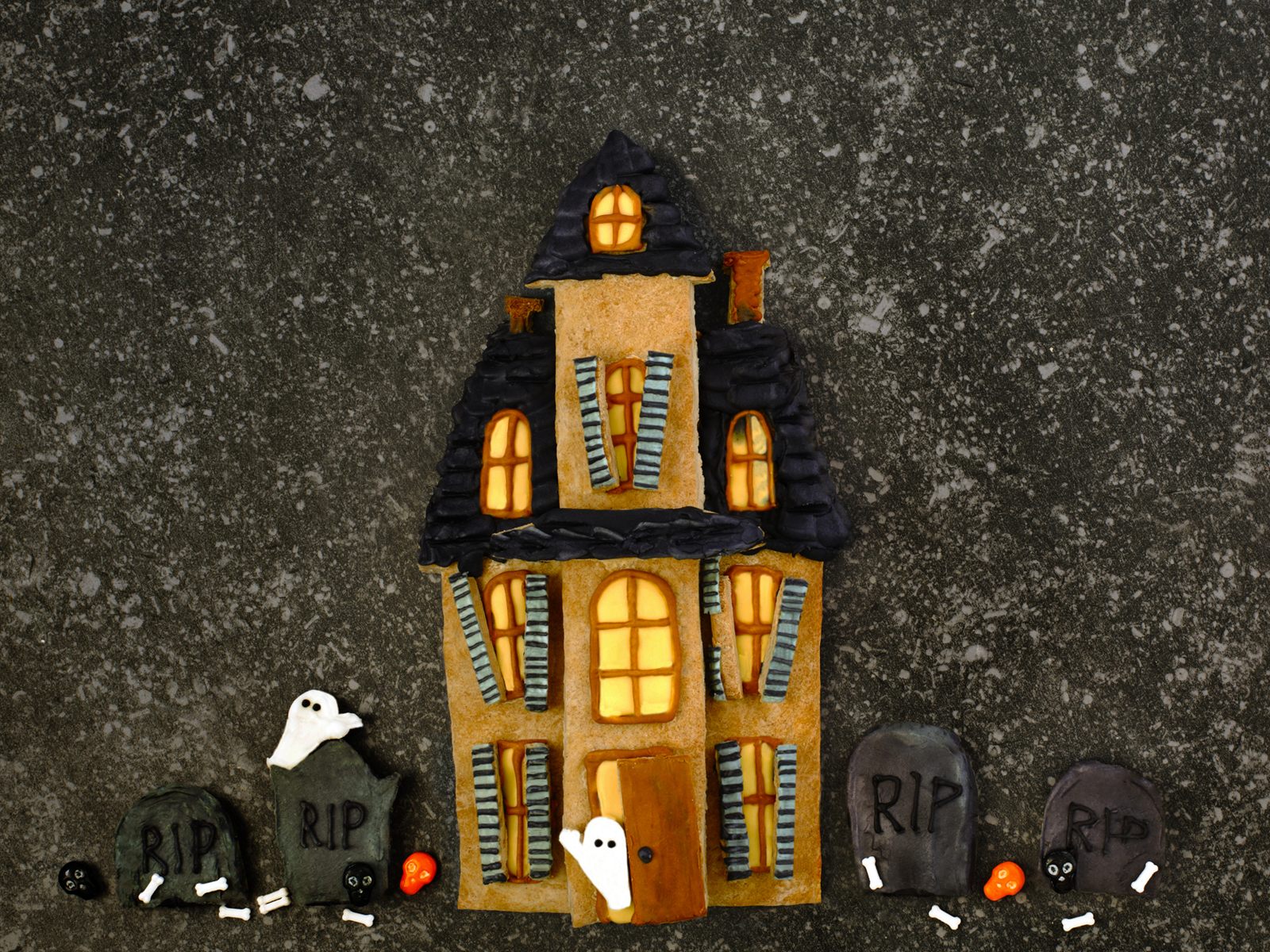 tall victorian gingerbread haunted house with glowing amber windows, striped shutters and a row of rip tombstones on dark stone