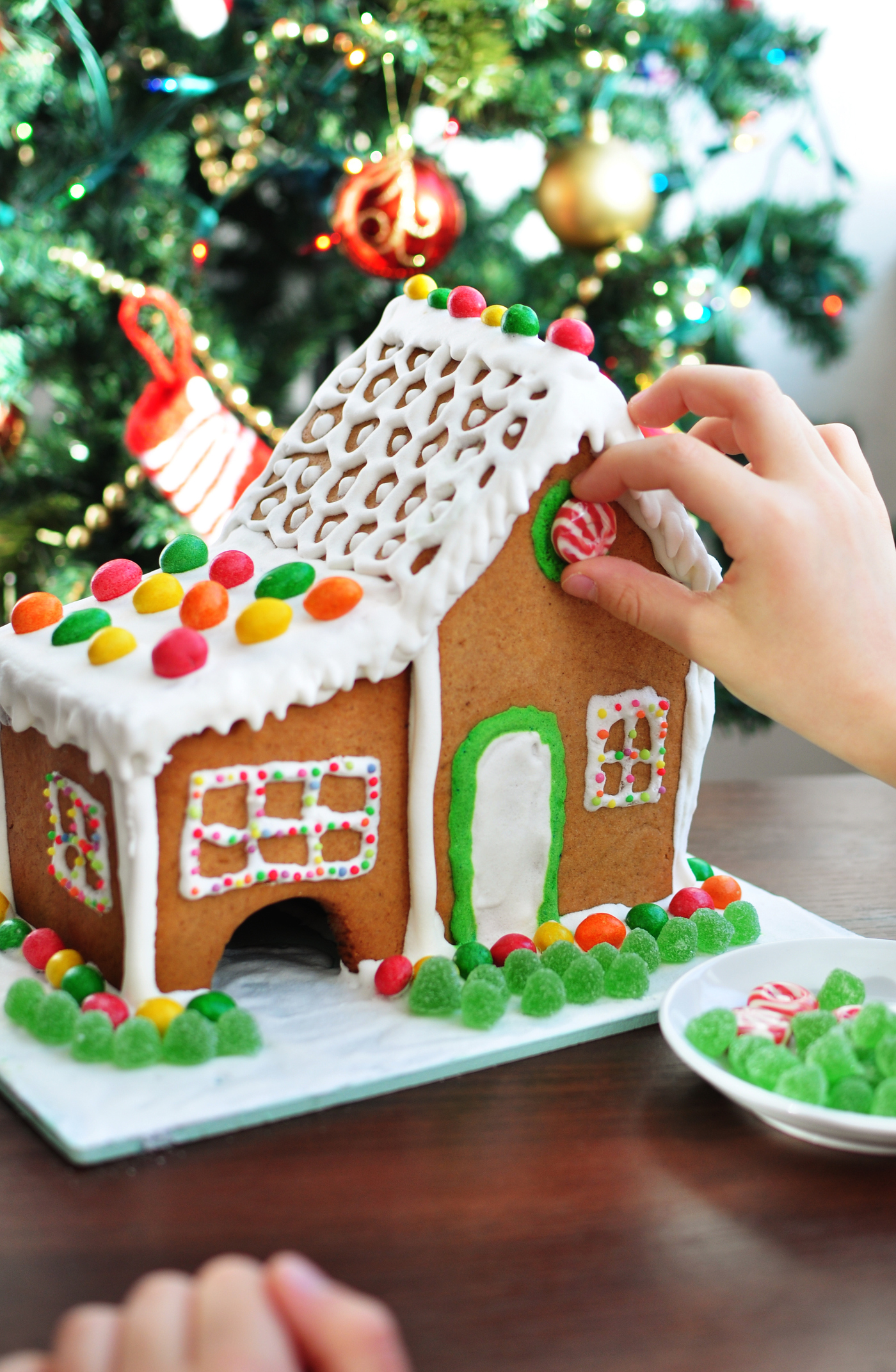 child's hand pressing a peppermint candy onto a white iced gingerbread house in front of a christmas tree