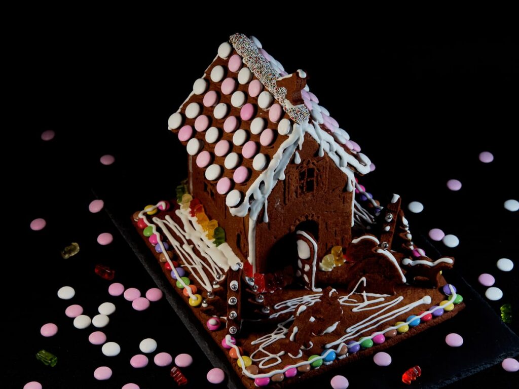 gingerbread cottage with a candy button roof and a candy border along the base, with candy buttons and gummy bears scattered on the dark board around it
