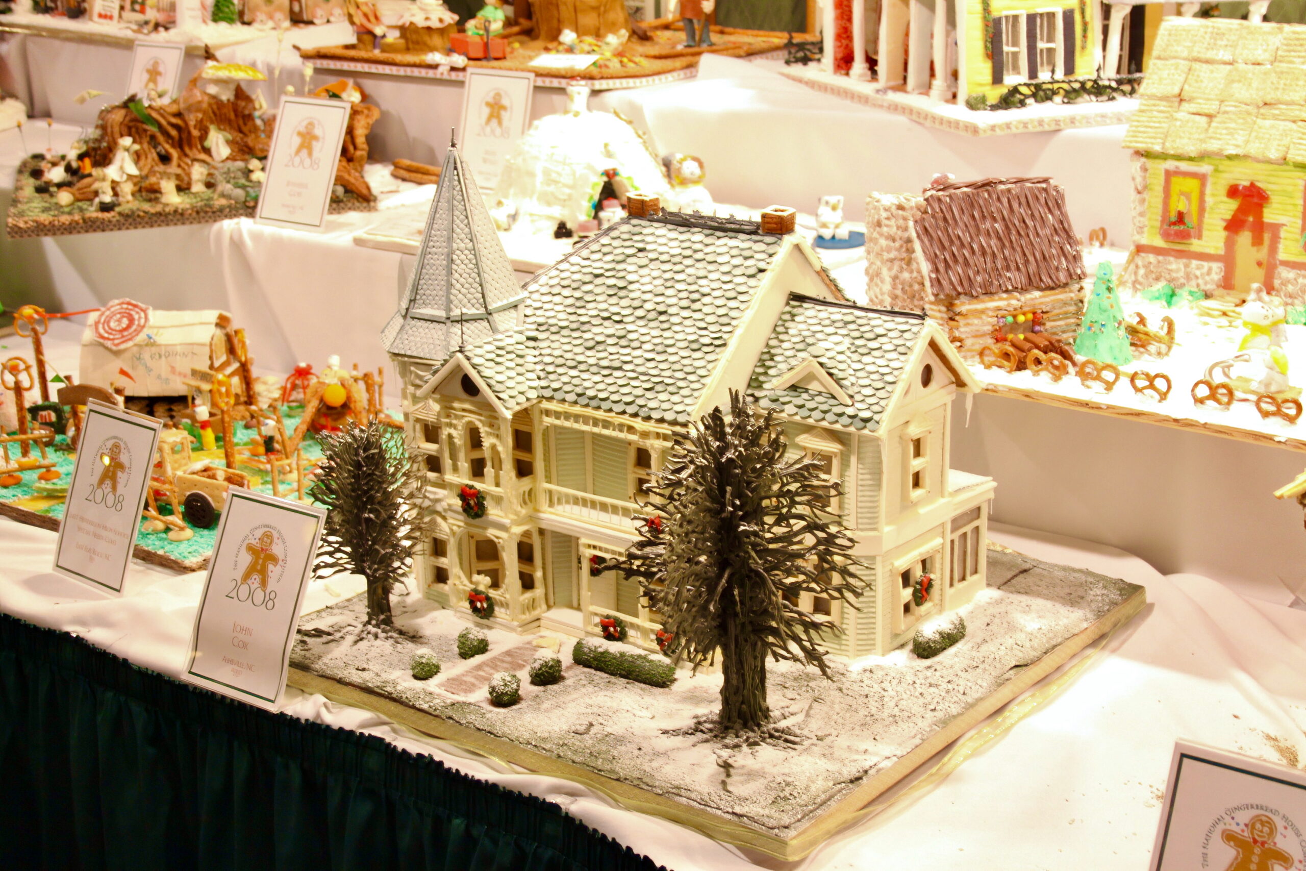 Rows of small competition gingerbread houses on white-draped tables, a pale Victorian house in front